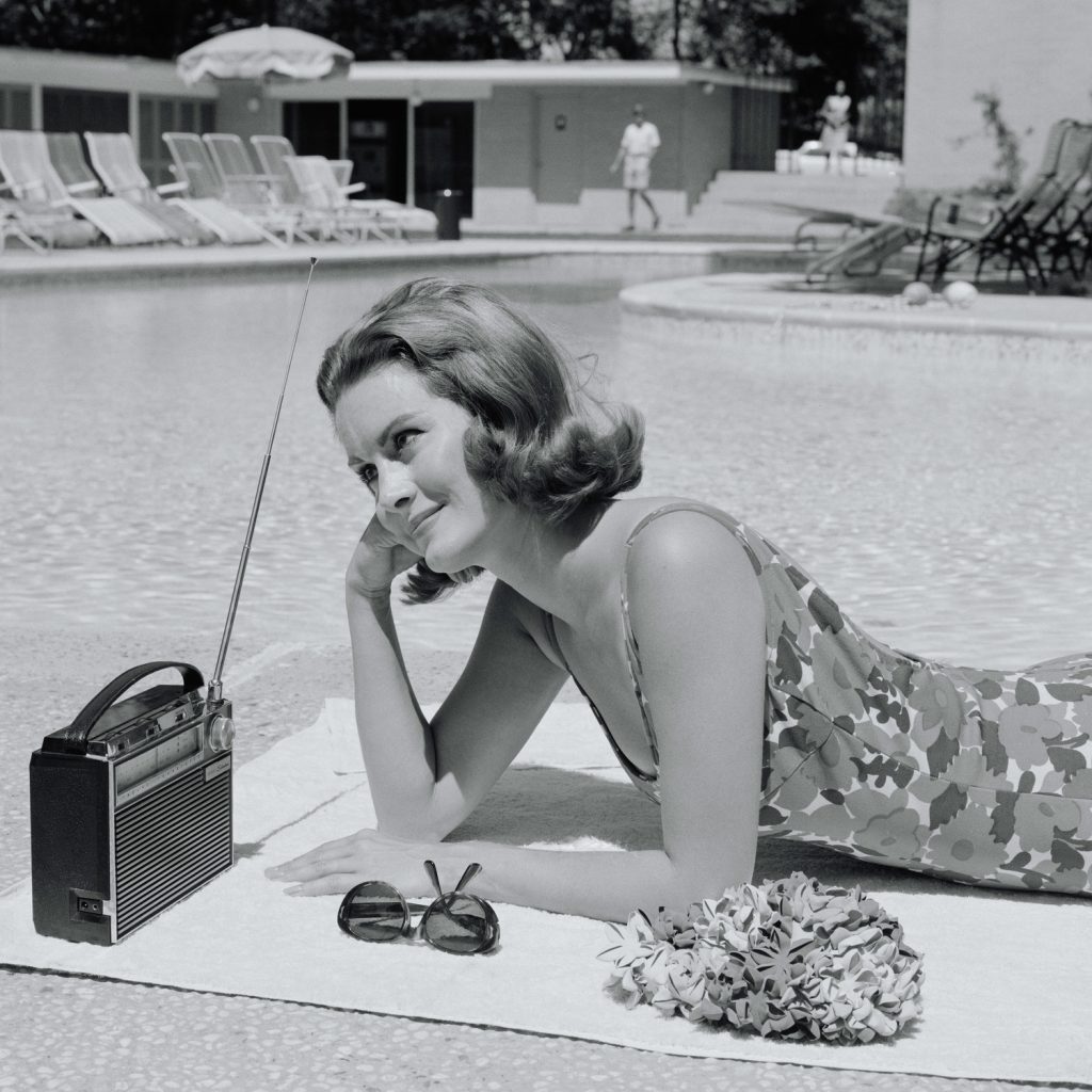 UNITED STATES - CIRCA 1960s:  Woman lying on towel poolside listening to radio with antenna up.